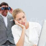 A thoughtful woman stares at a computer screen, propping her head up with her hand. Behind her back, a spy with binoculars put to his eyes.