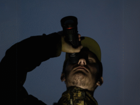 A man wearing a cap is holding a monocular and looking through it into the dark night, with only darkness visible in the background.